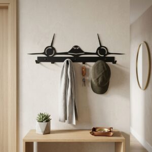 Czarny metalowy wieszak ścienny w kształcie samolotu SR-71 Blackbird z zawieszoną szarą chustą i oliwkową czapką z daszkiem, zamontowany na jasnej ścianie nad drewnianą konsolą.