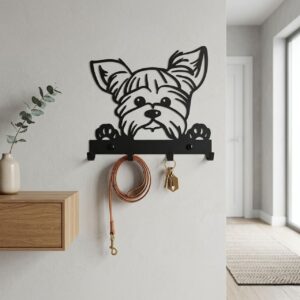 Czarna, metalowa dekoracja ścienna w kształcie głowy psa rasy Yorkshire Terrier z łapkami, pełniąca funkcję wieszaka. Zamontowana na jasnej, szarej ścianie. Na haczykach wisi zwinięta, skórzana smycz oraz pęk kluczy.