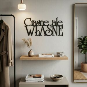 Czarny, metalowy napis Ciasne ale własne zawieszony na jasnej ścianie w nowoczesnym, niewielkim, ale stylowo urządzonym salonie nad jasną kanapą.