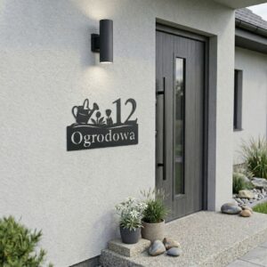 Metalowa tabliczka adresowa z numerem domu i wzorem konewki z kwiatami, zamontowana na jasnej elewacji obok drzwi wejściowych