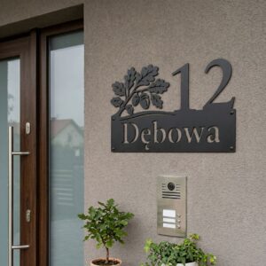 Metalowa tabliczka adresowa na dom z wyciętym numerem, nazwą ulicy i motywem liści dębu, zamontowana na szarej elewacji