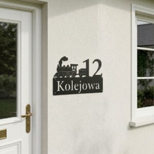 Czarna metalowa tabliczka na dom z numerem 12, napisem Kolejowa i motywem pociągu, zamontowana na jasnej ścianie budynku.