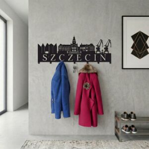 Czarny, metalowy wieszak ścienny z wyciętymi zabytkami Szczecina i napisem, zawieszony na jasnej ścianie z powieszonymi ubraniami.