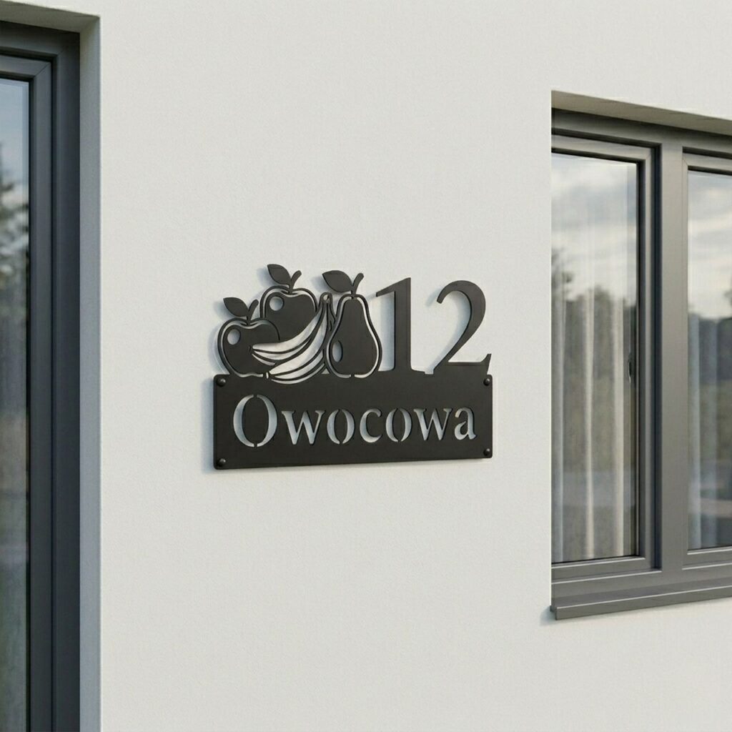 Nowoczesna czarna tabliczka adresowa z wyciętym laserowo motywem owoców (jabłka, banany, gruszka) oraz adresem ulica Owocowa 12, zamontowana na białej ścianie domu pomiędzy oknami.