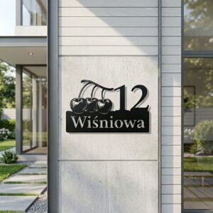 Czarna metalowa tabliczka adresowa z wyciętym motywem trzech wisienek i adresem ulica Wiśniowa 12, zamontowana na jasnym filarze nowoczesnego budynku z dużymi oknami.