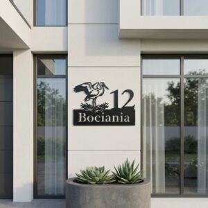 Personalizowana metalowa tabliczka adresowa na dom z numerem i motywem bociana zamontowana na jasnej elewacji.