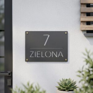 Szara metalowa tabliczka z numerem 7 i nazwą ulicy Zielona zamontowana na białej elewacji budynku