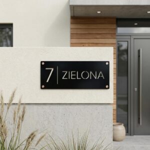 Czarna pozioma tabliczka z numerem 7 i ulicą Zielona zamontowana na betonowym murku ogrodzeniowym z ozdobnymi zaślepkami