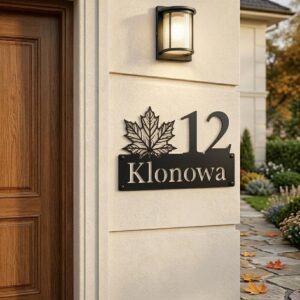 Metalowa tabliczka adresowa z motywem liścia klonu na jasnym tynku obok lampy i drewnianych drzwi
